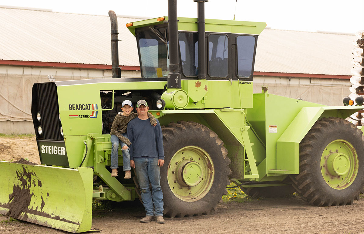 Farmer and son standing next to farming equipment
